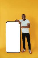 Leaning on, the large ,smartphone for copy space. Black man is in the studio against yellow background photo