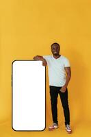 Leaning, on,, the large smartphone for copy space. Black man is in the studio against yellow background photo