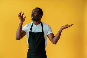 Bartender in uniform is showing ,delicious,gestures. Black man is in the studio against yellow background photo