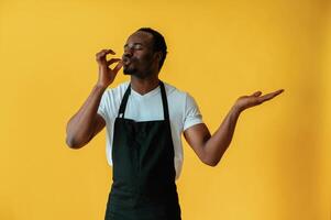 Bartender in ,uniform is showing delicious gestures. Black man is in the studio against yellow background photo