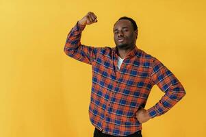 Showing muscles, hand gesture. Black man is in the studio against yellow background photo