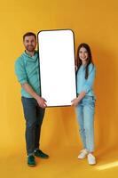 Holding ,white display, of smartphone, copy space. Man and woman are standing against yellow background photo