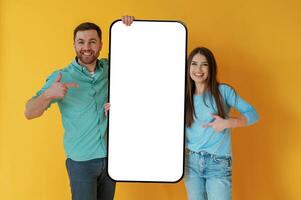 Holding white,, display of smartphone, copy space. Man and woman are standing against yellow background photo
