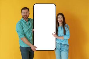 Display for copy space. Man and woman are standing with large smartphone against yellow background photo