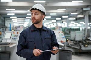 Looking far away, holding notepad.,, Factory worker is indoors with hard hat photo