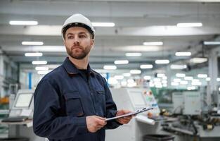 Looking far away, holding ,notepad. Factory, worker is indoors with hard hat photo