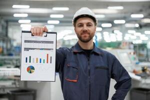 Plan with graphs, in, hand. Factory worker is indoors with hard hat photo