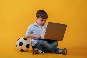 Using, laptop. Soccer ball on the floor.. Little boy is in the studio against yellow background photo