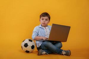 Using, laptop. Soccer, ball on the floor. Little boy is in the studio against yellow background photo