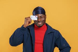 Covering eye by credit card. Handsome black man is in the studio against yellow background, photo