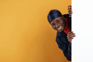 White copy space, smiling and hiding behind it. Handsome black man is in the studio against yellow background, photo