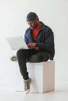 Sitting and using laptop. Black man in cap is in the studio against white background, photo