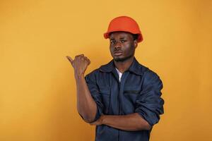 Guy in hard hat, showing ,copy,, space by finger. Handsome black man is in the studio against yellow background photo