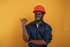 Guy, in ,hard, hat showing copy space by finger. Handsome black man is in the studio against yellow background photo