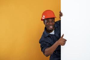Engineer in hard, hat is,, hiding behind white copy space. Handsome black man is in the studio against yellow background photo