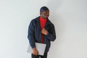 Standing ,and ,holding laptop ,in hand. Black man in cap is in the studio against white background photo