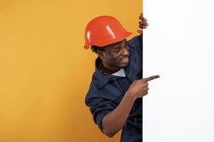 Engineer, in ,hard hat is hiding, behind white copy space. Handsome black man is in the studio against yellow background photo