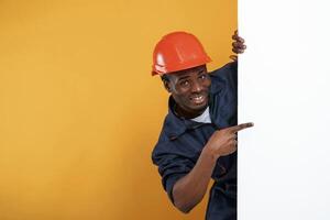 Engineer in hard hat is hiding behind, white copy space. Handsome black man is in the studio against yellow background photo