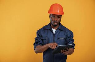 Digital tablet in hand, construction,, worker. Handsome black man is in the studio against yellow background photo