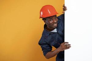 Engineer, in hard hat is hiding behind white copy space. Handsome black man is in the studio against yellow background photo