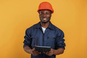 Construction, worker in hard, hat and with tablet. Handsome black man is in the studio against yellow background photo