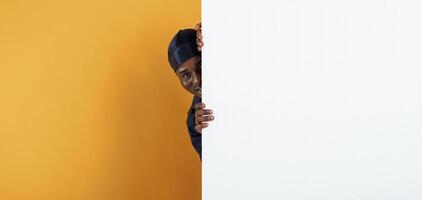 White, copy space, smiling and hiding behind it. Handsome black man is in the studio against yellow background photo