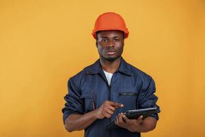Digital tablet in hand, construction, worker. Handsome black man is in the studio against yellow background photo