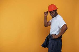 Standing and posing,, construction worker in hard hat. Handsome black man is in the studio against yellow background photo