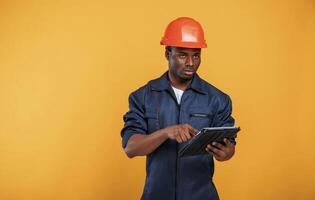Construction worker, in hard hat and with tablet. Handsome black man is in the studio against yellow background photo