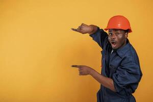 Guy in hard, hat showing copy space by fingers. Handsome black man is in the studio against yellow background photo