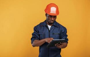 Construction, worker in hard hat and with tablet. Handsome black man is in the studio against yellow background photo