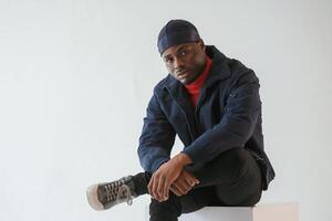 Sitting on the cube ,chair. Black man in cap is in the studio against white background photo