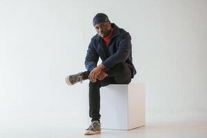 Sitting on the ,cube chair. Black man in cap is in the studio against white background photo