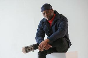 Sitting on ,the cube chair. Black man in cap is in the studio against white background photo