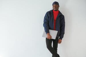 Standing and, holding laptop in hand. Black man in cap is in the studio against white background photo