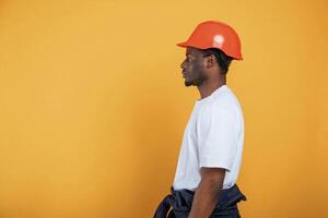 Standing and ,,posing, construction worker in hard hat. Handsome black man is in the studio against yellow background photo