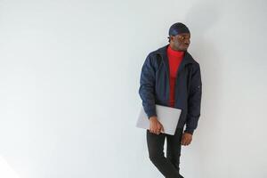 Standing, and holding laptop in hand. Black man in cap is in the studio against white background photo