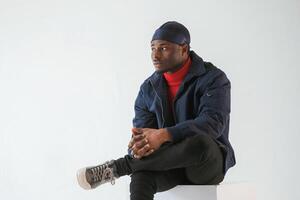 Sitting, on the cube chair. Black man in cap is in the studio against white background photo