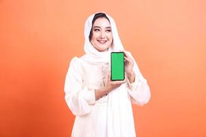 A smiling Muslim woman in a hijab is holding a smartphone with a green screen. She is looking away from the camera. isolated on a orange background photo