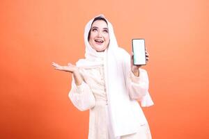 A happy muslim woman in hijab presenting a mobile phone app. She is smiling and looking upwards while holding a smartphone. isolated on a orange background. photo