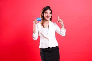 A woman in a white suit holds a credit card and points up with her index finger while smiling, isolated on a red background photo