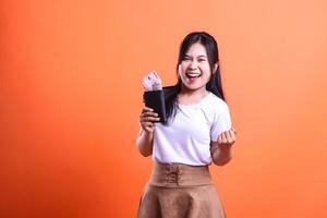 A young woman smiling broadly while holding a black wallet containing rupiah money in her hand isolated on an orange background wearing a white shirt and brown skirt and very happy. photo