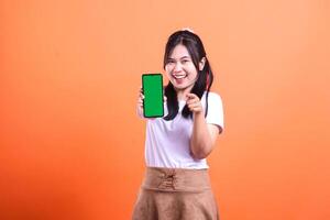 Smiling woman holding a smartphone with green screen, on orange background, and pointing at camera photo
