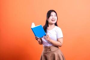 A woman in a white shirt and brown skirt holds a blue book and looks to the side isolated on orange background photo