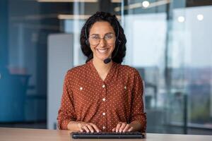 Portrait of young successful Latin American online support worker, woman smiling and looking at camera with headset phone, webcam view, typing on keyboard, inside office at workplace. photo