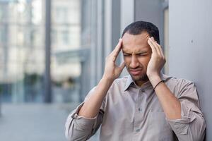 Portrait of adult man in shirt leaning on building wall while keeping both hands on temples with closed eyes. Sick guy suffering from migraine attack and massaging head fro relief on fresh air. photo