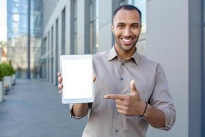 Portrait of a smiling young hispanic man standing near an office building, holding a mock-up tablet and pointing at a white screen. photo