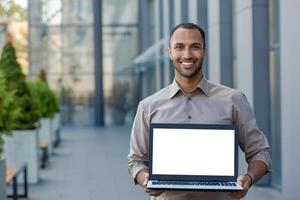 Portrait of a smiling hispanic man holding a mockup of a laptop with a white screen, standing on the street near an office building and showing the device to the camera. photo