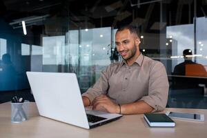 Young successful businessman working inside the office with a laptop, satisfied with the results of the achievement, the man is smiling, typing on the keyboard. photo
