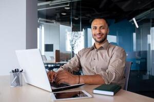 Attractive african american man sitting at desk in front of laptop in office, programmer working in modern environment, tapping on keyboard and smiling at camera. photo